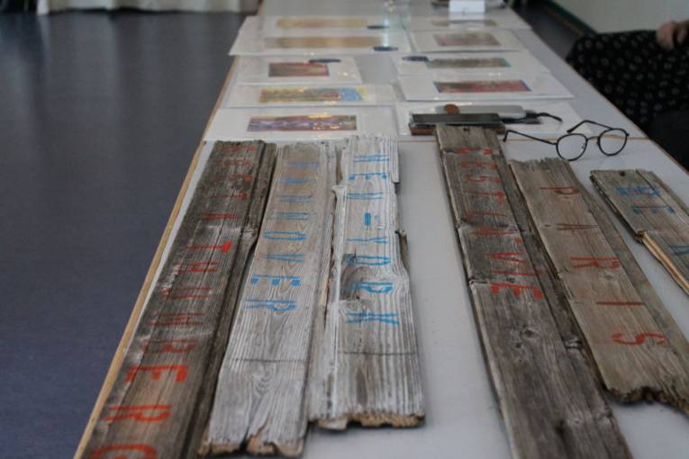 Hier sieht man alte Holzlatten, die mit Worten bedruckt sind. Im Hintergrund liegen kleine Kunstdrucke auf einem Tisch.